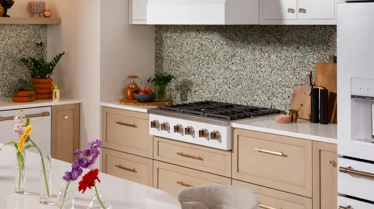 Modern kitchen with white cabinets above a colorful confetti backsplash and natural wooden drawers below with a built in gas stove featuring other white appliances with gold accents.